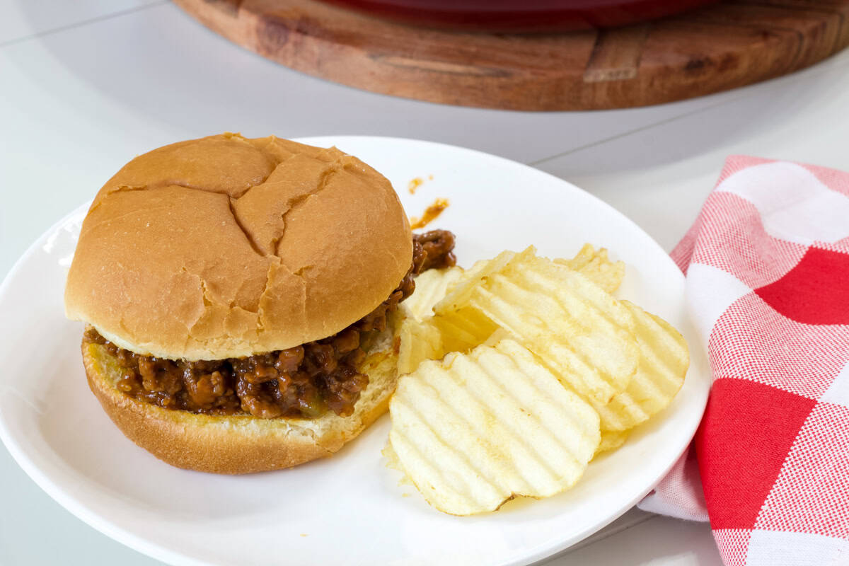 One sloppy joe sandwich on a white plate with come potato chips next to it.