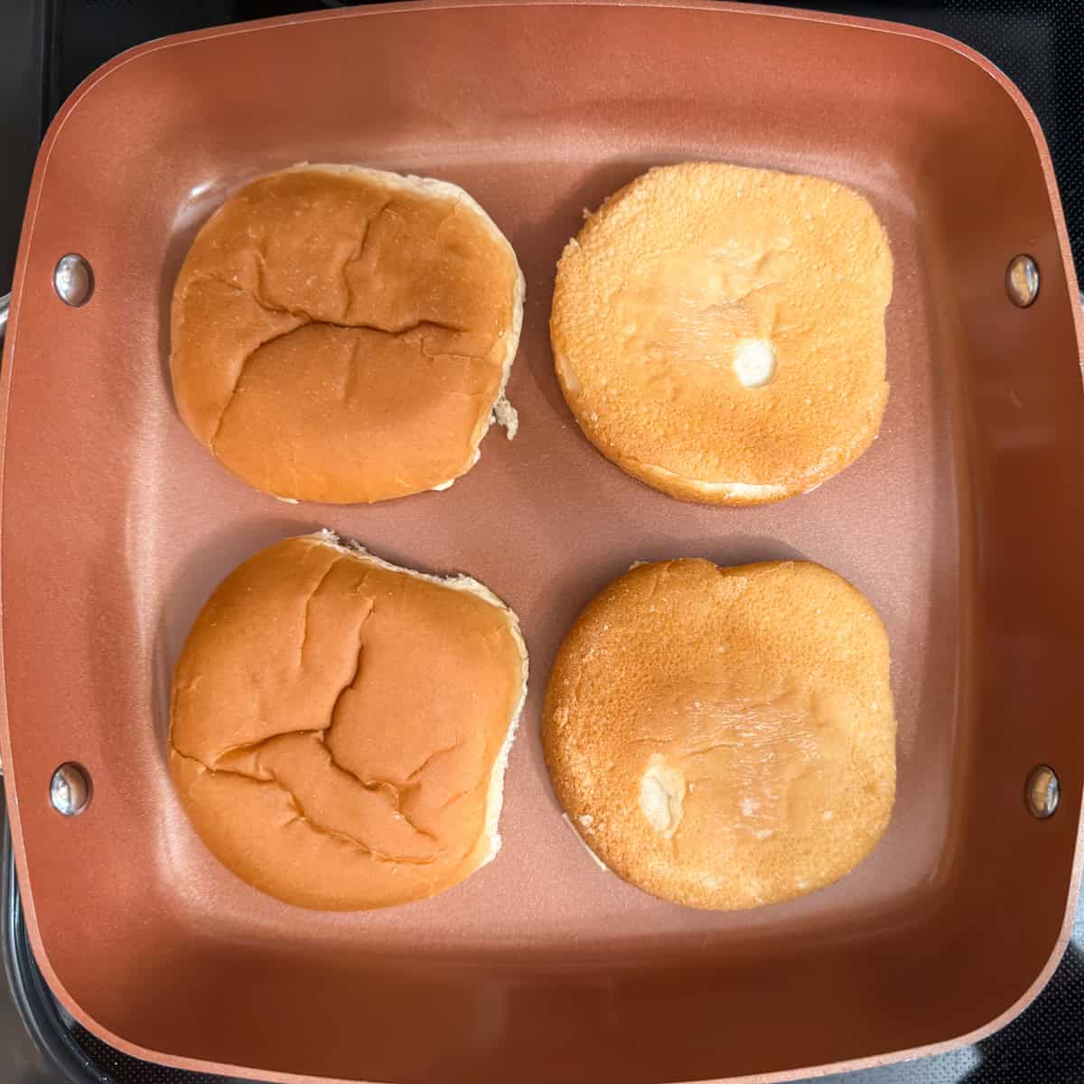 Four buttered bun halves face down in a square skillet being toasted.