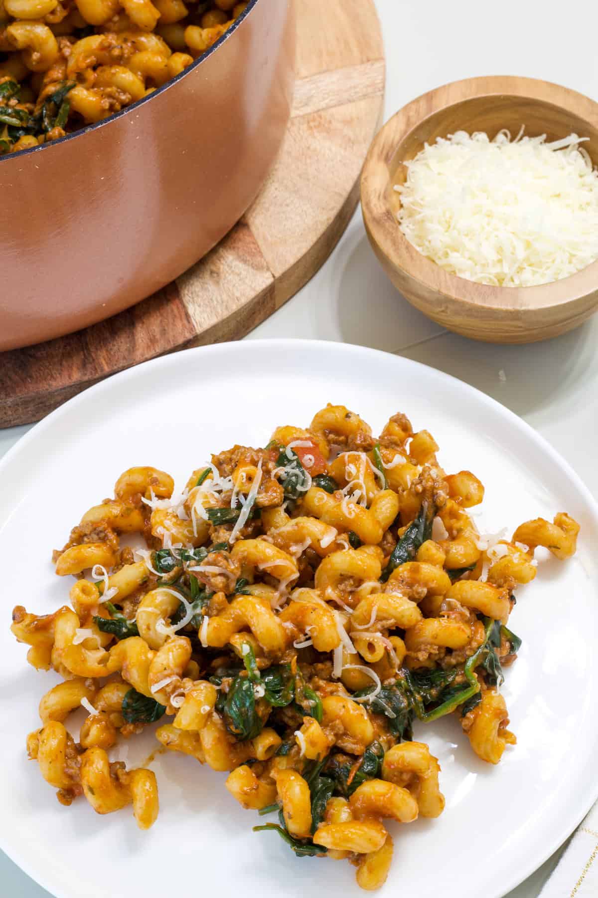 A plate of pasta with cheese and spinach, with a bowl of grated parmesan cheese in the background.