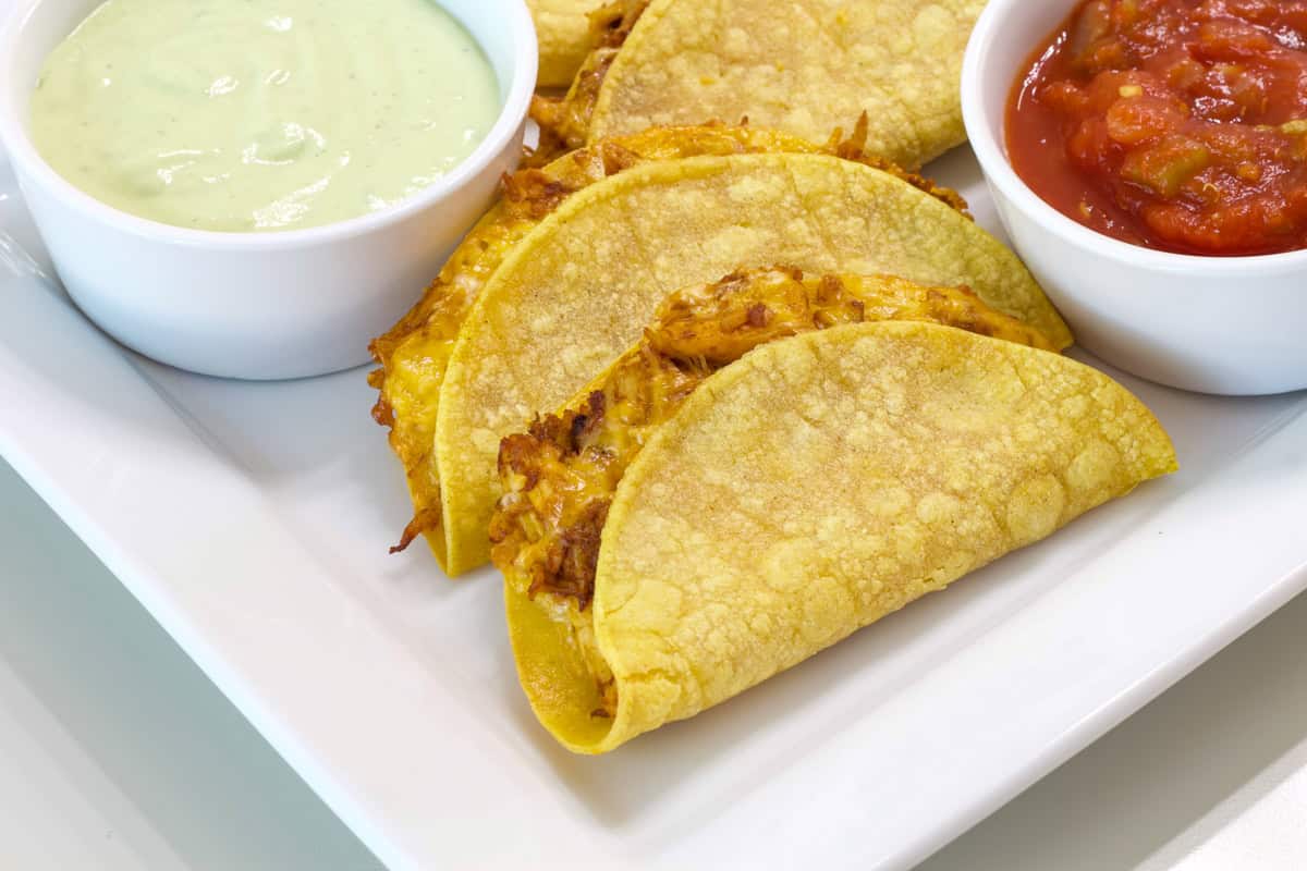 Several baked tacos on a white plate with a small bowl of dipping sauce and a small bowl of salsa.