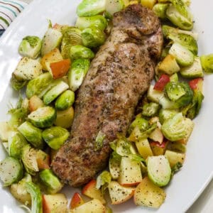 The uncut pork tenderloin on a platter surrounded by Brussels sprouts and apples.