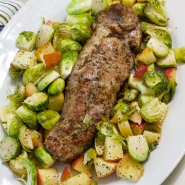 The uncut pork tenderloin on a platter surrounded by Brussels sprouts and apples.