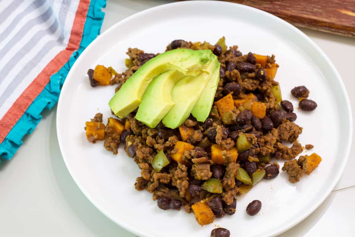 One serving of Ground Beef Sweet Potato Hash on a plate topped with three slices of avocado.